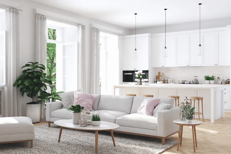 A bright, modern open-plan living room and kitchen with white furniture, light wood accents, potted plants, large windows, and pendant lights. Sunlight streams in, creating an airy, inviting atmosphere.